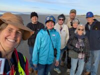 Smiling woman with hat taking selfie with six coastwatchers, dressed in layers, some with sunglasses, all smiling in the background on a sandy beach under blue skies